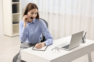 Teenager in headset taking notes while working at table indoors. Remote job