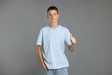 Photo of Teenage boy wearing light blue t-shirt and showing thumbs up on grey background