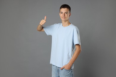 Photo of Teenage boy wearing light blue t-shirt and showing thumbs up on grey background