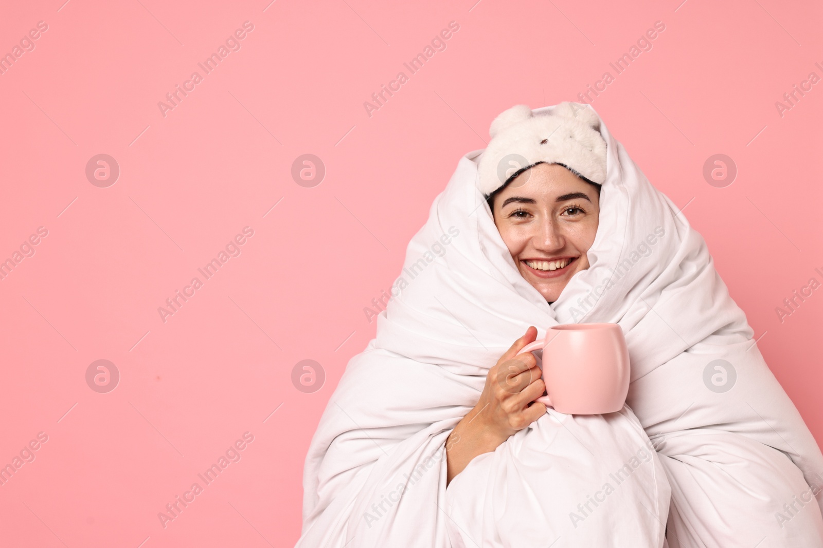 Photo of Happy young woman with cup wrapped in blanket on pink background. Space for text