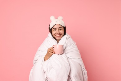 Happy young woman with cup wrapped in blanket on pink background