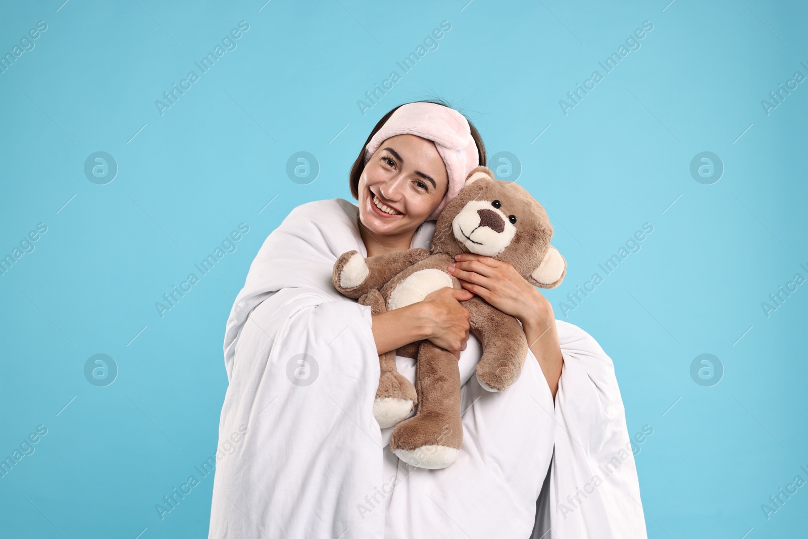 Photo of Overslept young woman with teddy bear wrapped in blanket on light blue background