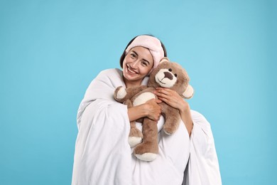 Overslept young woman with teddy bear wrapped in blanket on light blue background