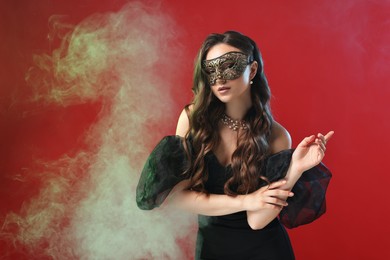 Photo of Beautiful woman wearing carnival mask on red background in color lights and smoke. Space for text