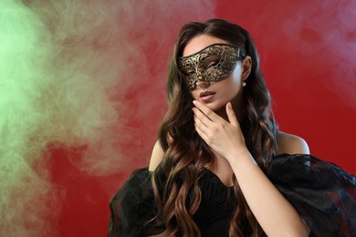 Beautiful woman wearing carnival mask on red background in color lights and smoke. Space for text