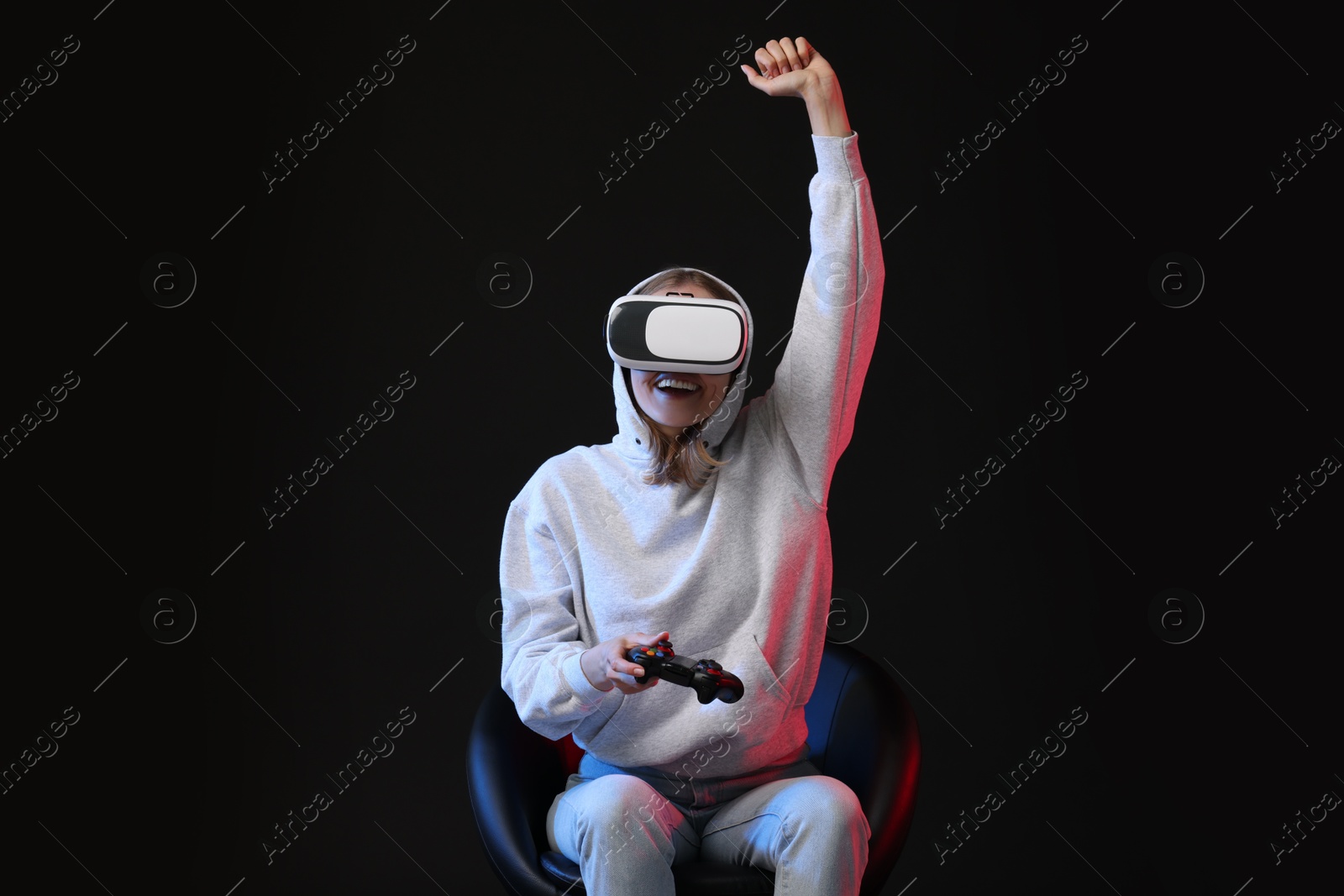 Photo of Happy woman with virtual reality headset and controller sitting on chair in neon lights against black background