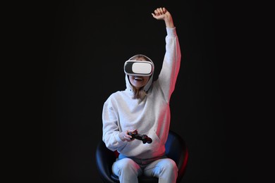 Happy woman with virtual reality headset and controller sitting on chair in neon lights against black background