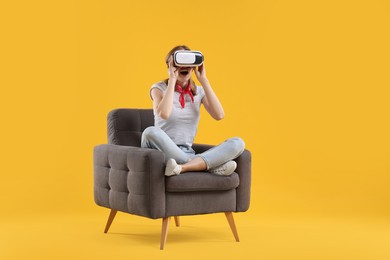 Emotional woman with virtual reality headset sitting in armchair on yellow background