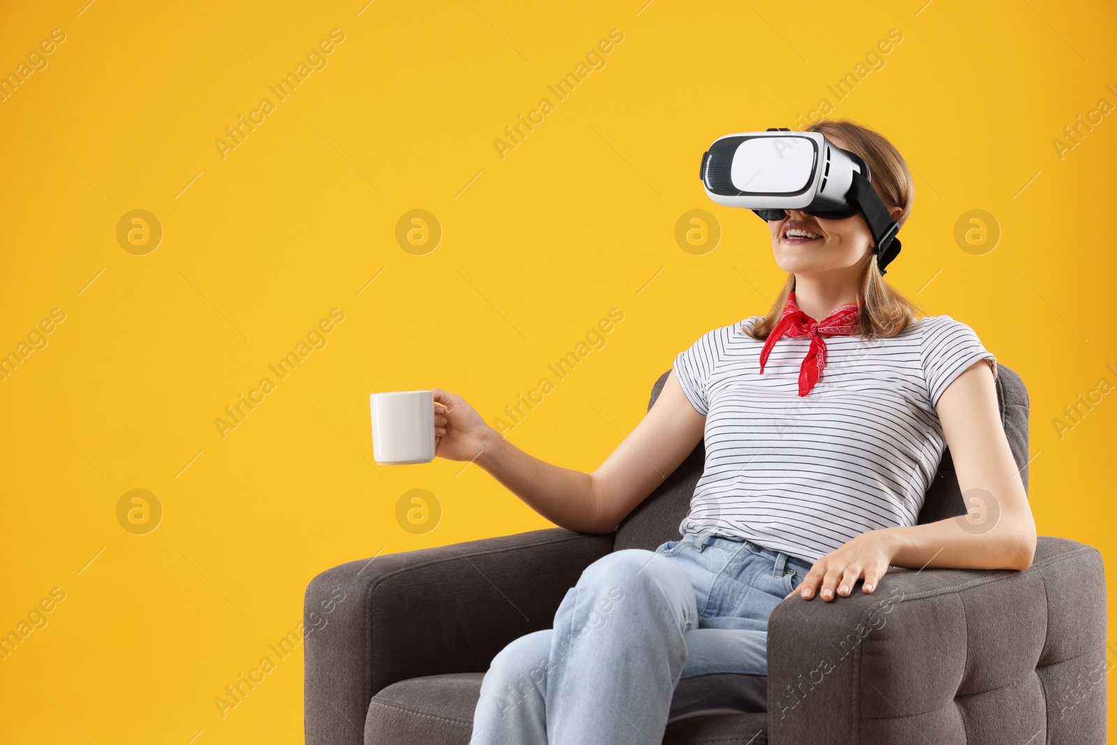 Photo of Happy woman with virtual reality headset and cup sitting in armchair on yellow background, space for text