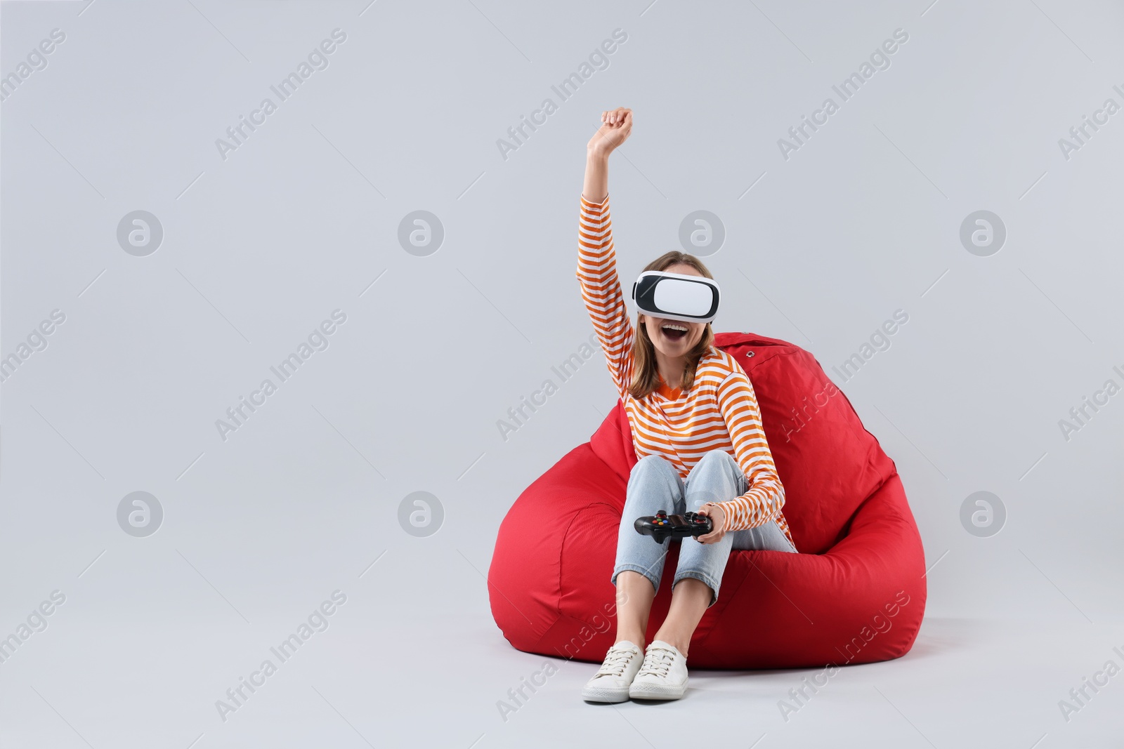 Photo of Happy woman with virtual reality headset and controller sitting on bean bag chair against gray background, space for text