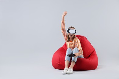 Photo of Happy woman with virtual reality headset and controller sitting on bean bag chair against gray background, space for text