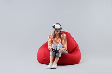 Photo of Happy woman with virtual reality headset and controller sitting on bean bag chair against gray background