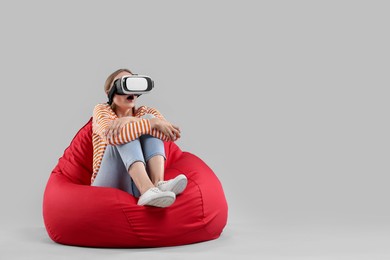 Emotional woman with virtual reality headset sitting on bean bag chair against gray background, space for text