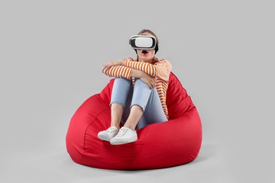 Emotional woman with virtual reality headset sitting on bean bag chair against gray background