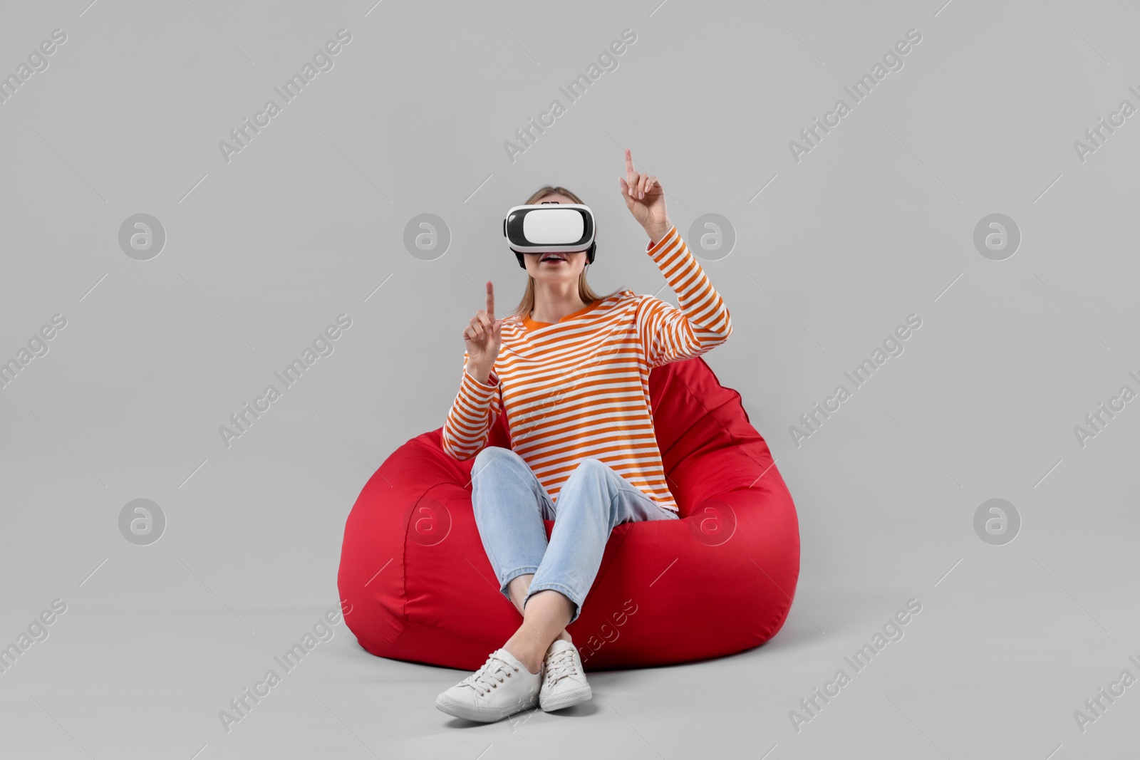 Photo of Happy woman with virtual reality headset sitting on bean bag chair against gray background