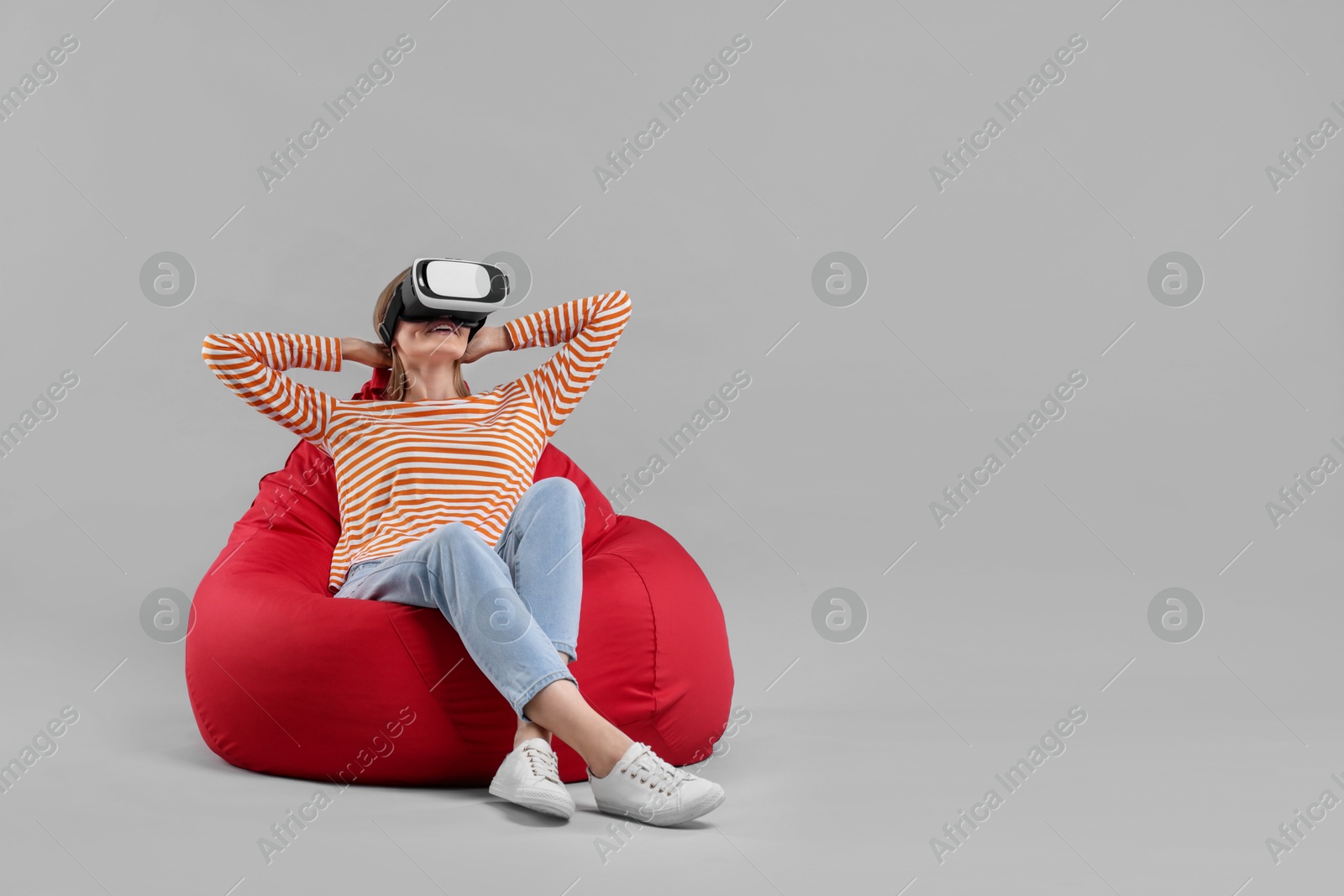 Photo of Happy woman with virtual reality headset sitting on bean bag chair against gray background, space for text