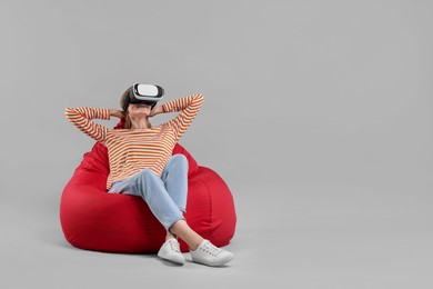 Happy woman with virtual reality headset sitting on bean bag chair against gray background, space for text