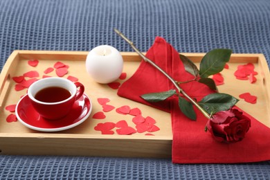 Wooden tray with burning candle, rose, cup of tea and red paper hearts on bed indoors