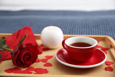 Wooden tray with burning candle, rose, cup of tea and red paper hearts on bed indoors, closeup