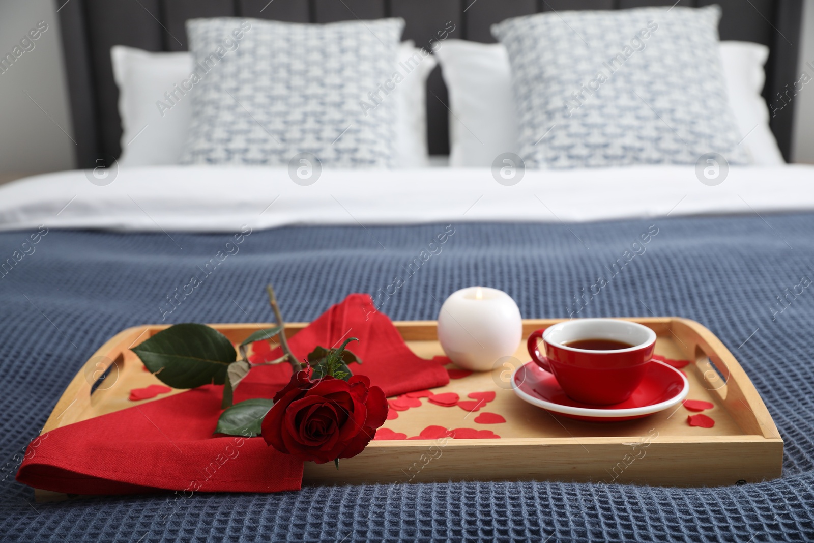 Photo of Wooden tray with burning candle, rose, cup of tea and red paper hearts on bed indoors