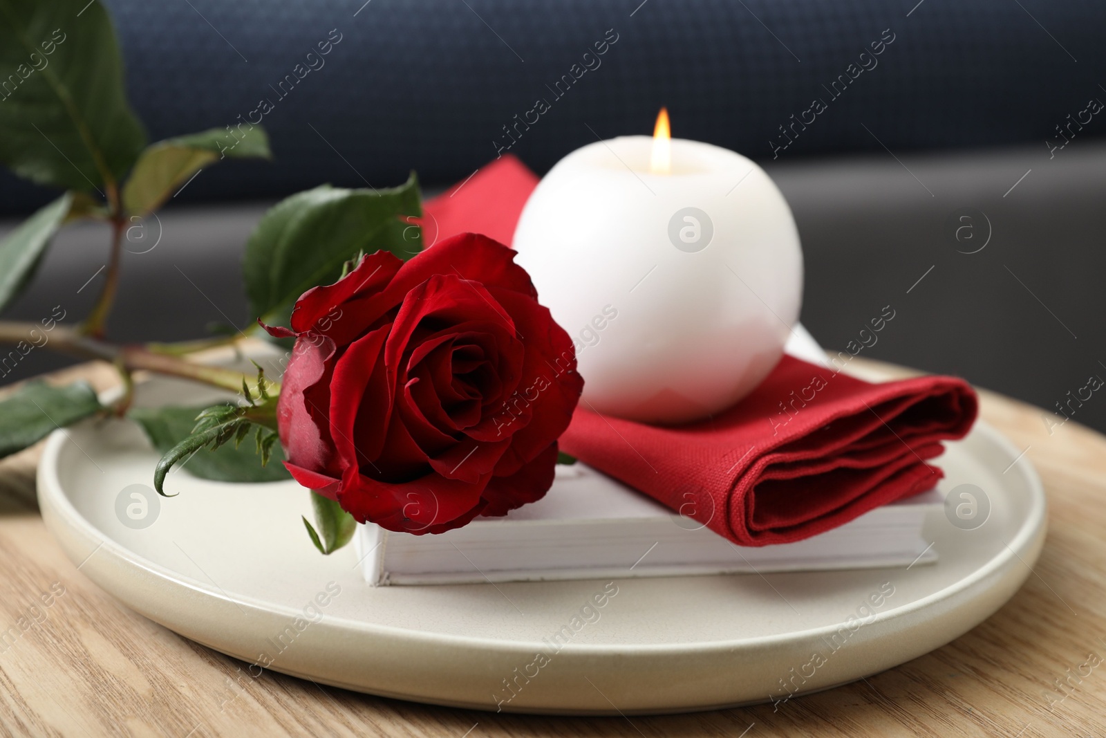 Photo of Burning candle, napkin, red rose and book on wooden table indoors, closeup