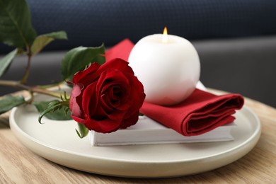 Photo of Burning candle, napkin, red rose and book on wooden table indoors, closeup