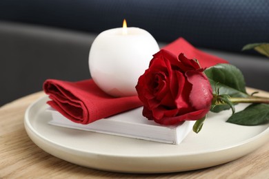 Photo of Burning candle, napkin, red rose and book on wooden table indoors, closeup