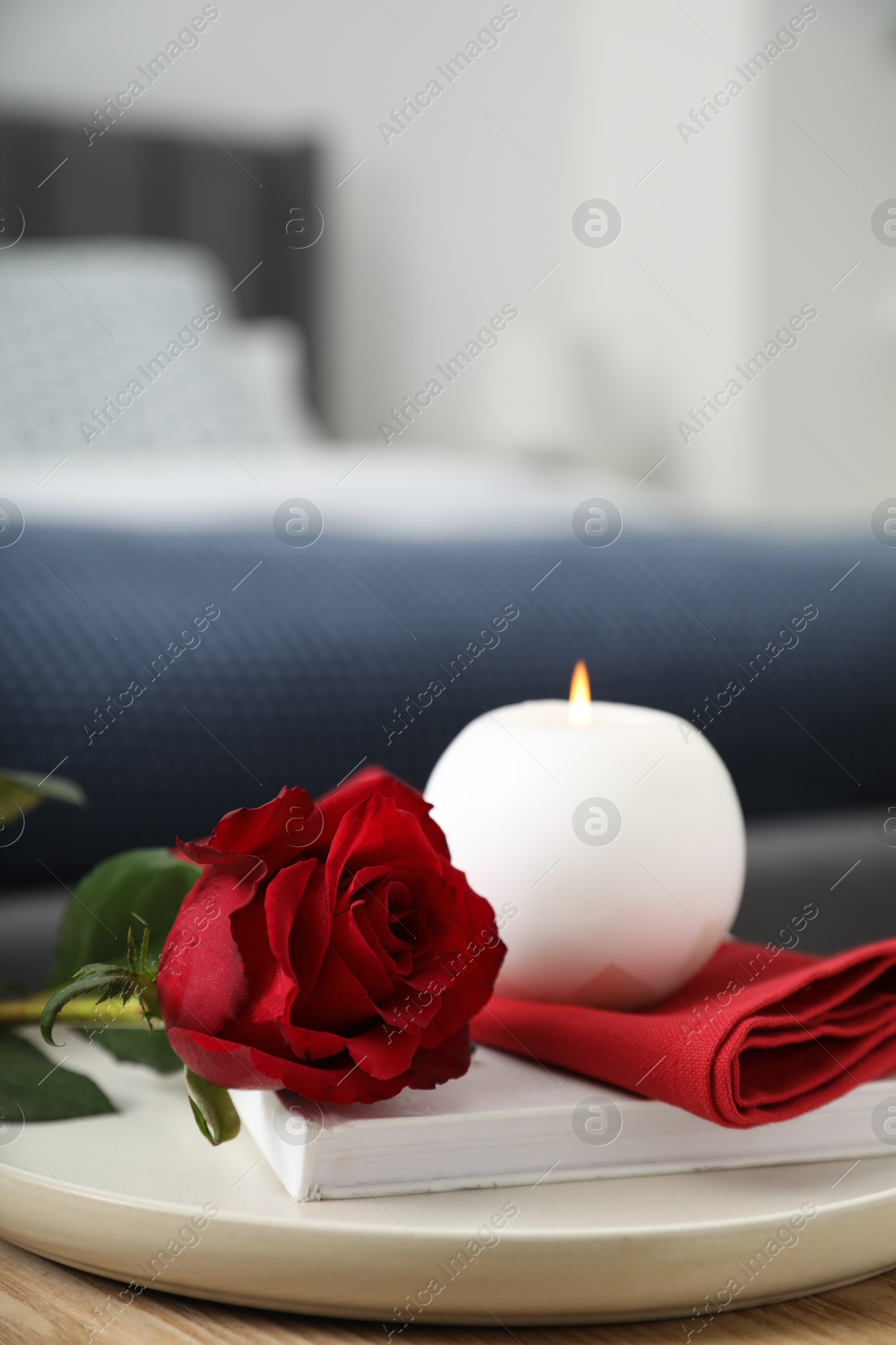 Photo of Burning candle, napkin, red rose and book on wooden table indoors, closeup