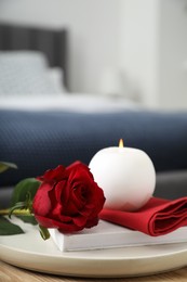 Photo of Burning candle, napkin, red rose and book on wooden table indoors, closeup