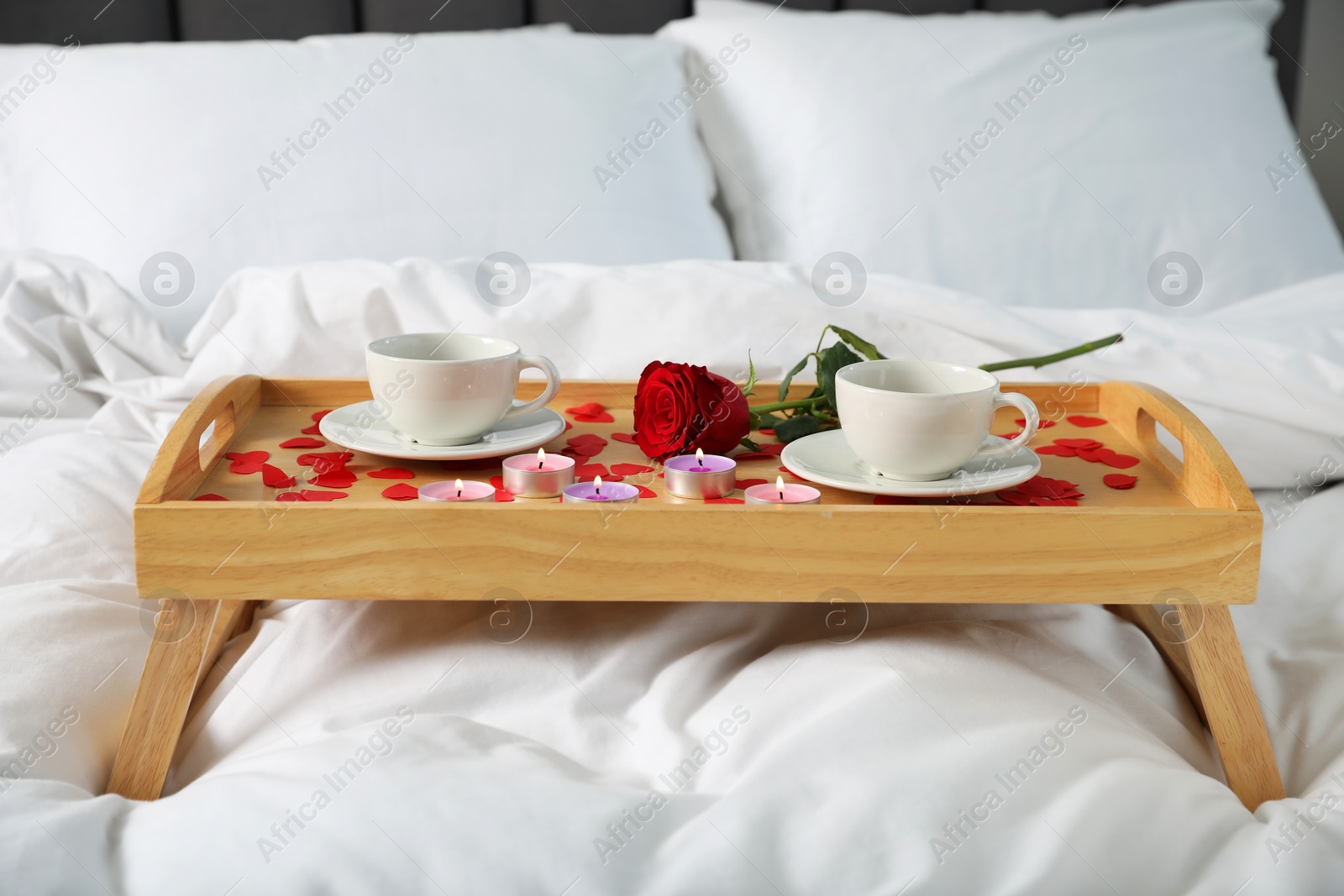 Photo of Wooden tray with burning candles, rose, cups of coffee and red paper hearts on bed indoors