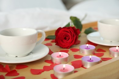 Wooden tray with burning candles, rose, cups of drink and red paper hearts on bed indoors, closeup