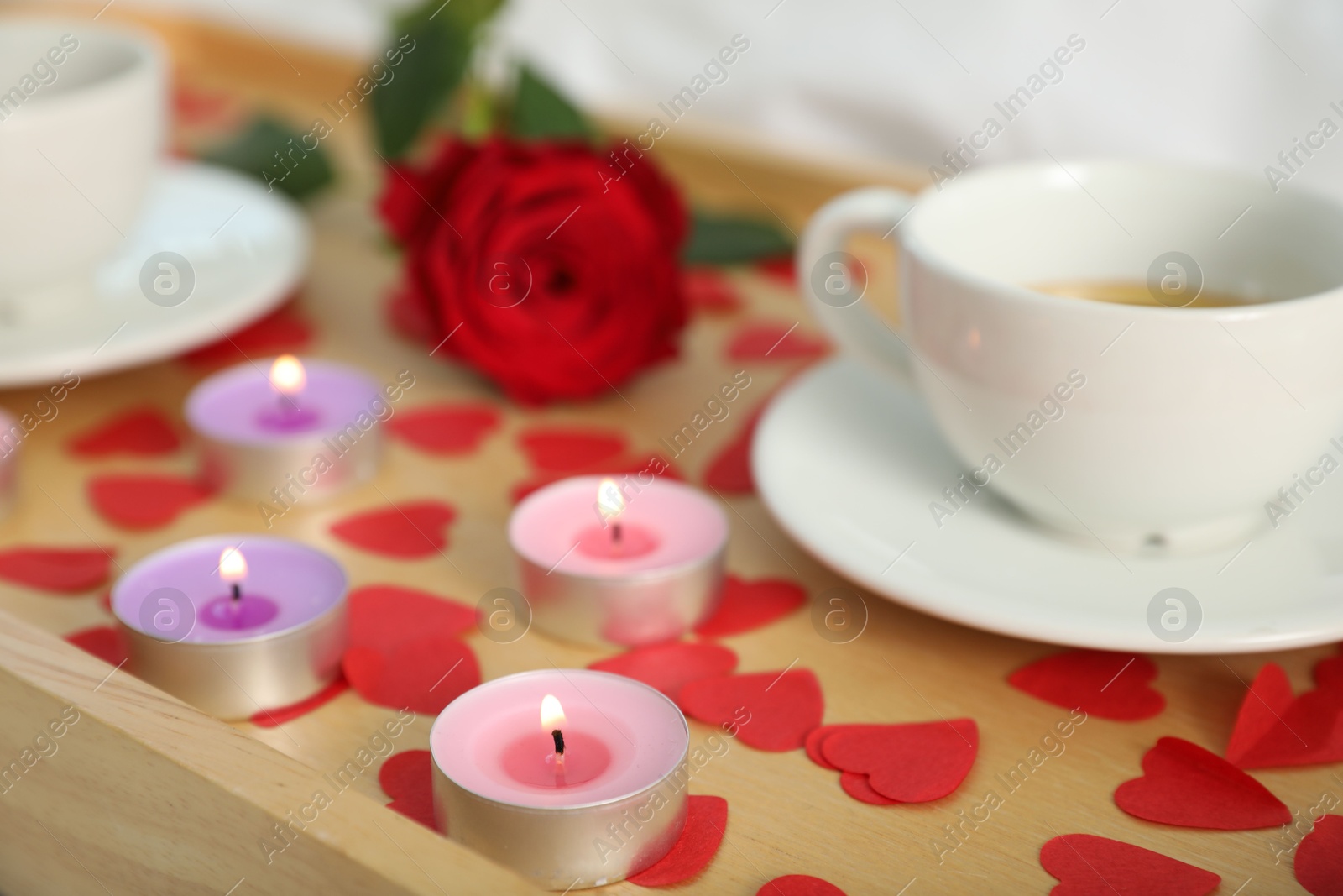 Photo of Wooden tray with burning candles, rose, cups of drink and red paper hearts on bed indoors, closeup