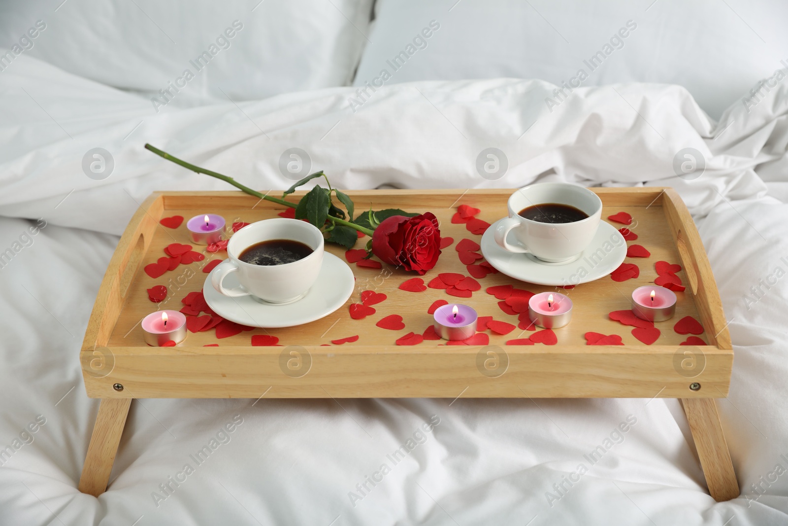 Photo of Wooden tray with burning candles, rose, cups of coffee and red paper hearts on bed indoors