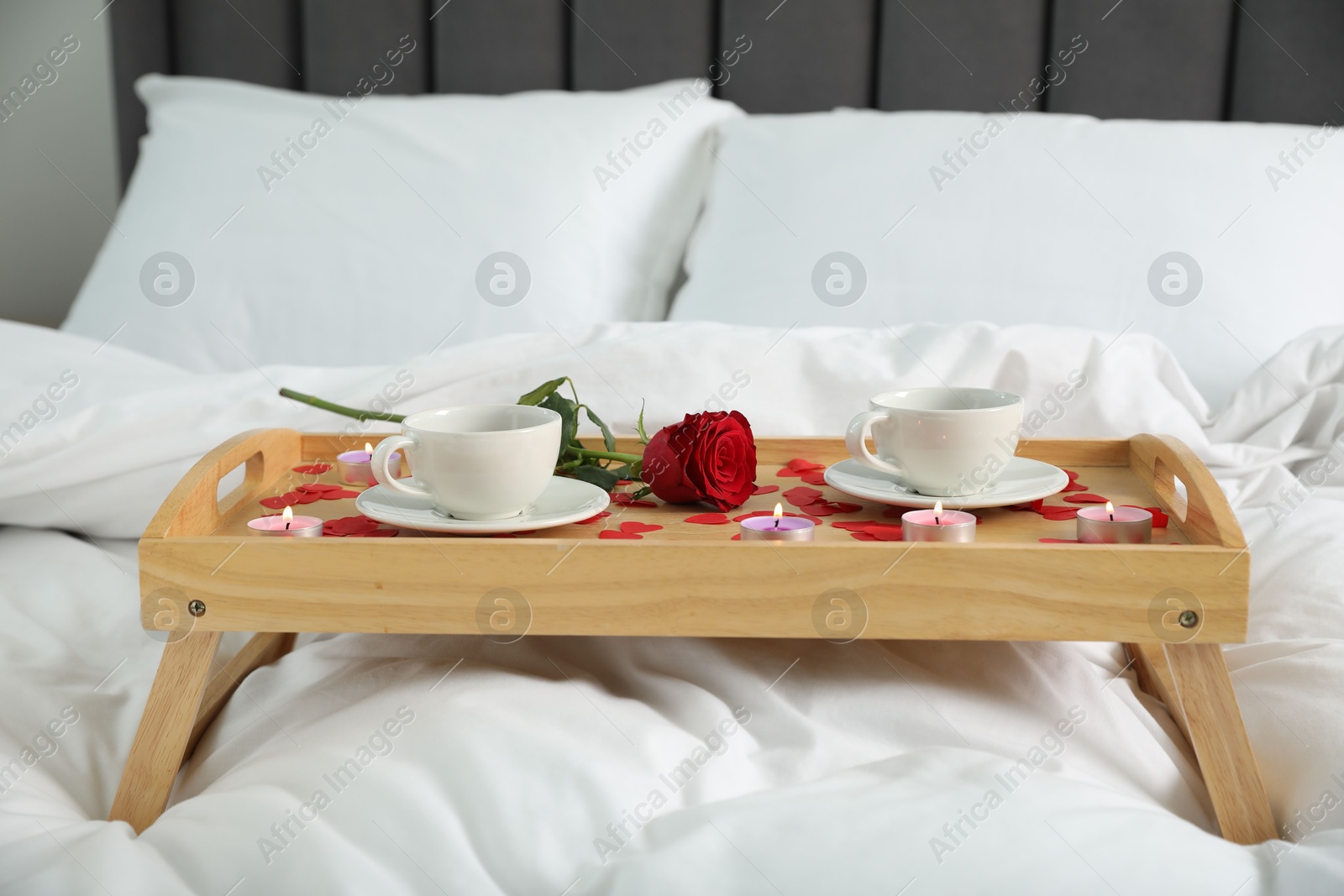 Photo of Wooden tray with burning candles, rose, cups of drink and red paper hearts on bed indoors