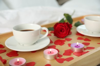 Photo of Wooden tray with burning candles, rose, cups of coffee and red paper hearts on bed indoors, closeup