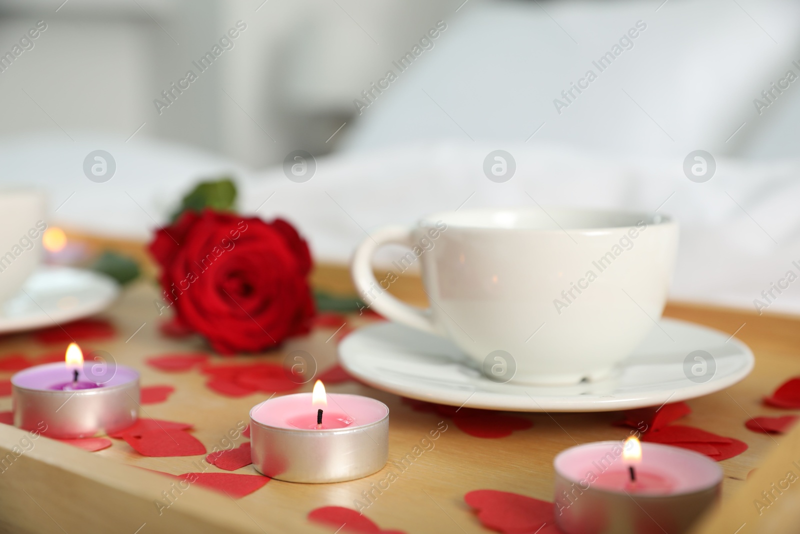 Photo of Wooden tray with burning candles, rose, cup of drink and red paper hearts on bed indoors, closeup