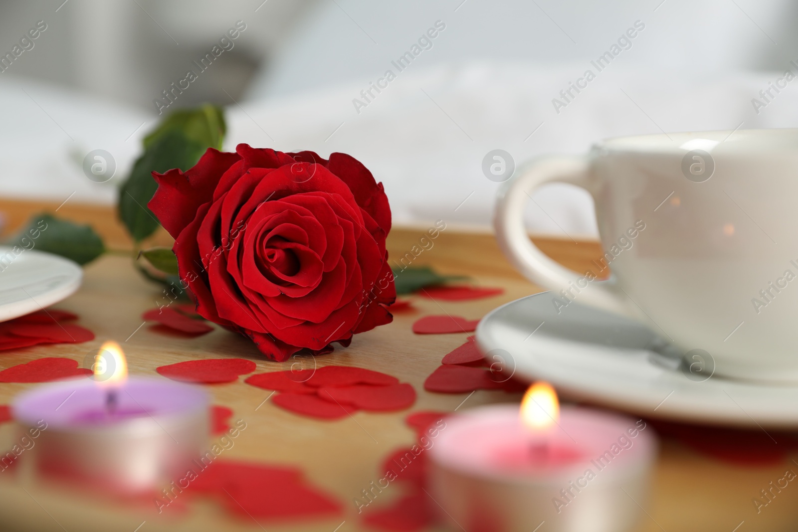 Photo of Wooden tray with burning candles, rose, cup of drink and red paper hearts on bed indoors, closeup