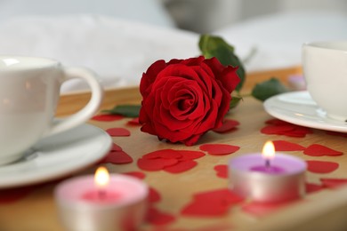 Photo of Wooden tray with burning candles, rose, cups of drink and red paper hearts on bed indoors, closeup