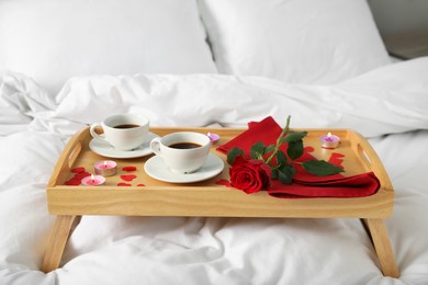 Photo of Wooden tray with burning candles, rose, cups of coffee and red paper hearts on bed indoors