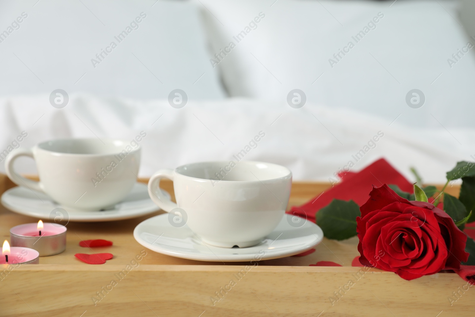 Photo of Wooden tray with burning candles, rose, cups of drink and red paper hearts on bed indoors