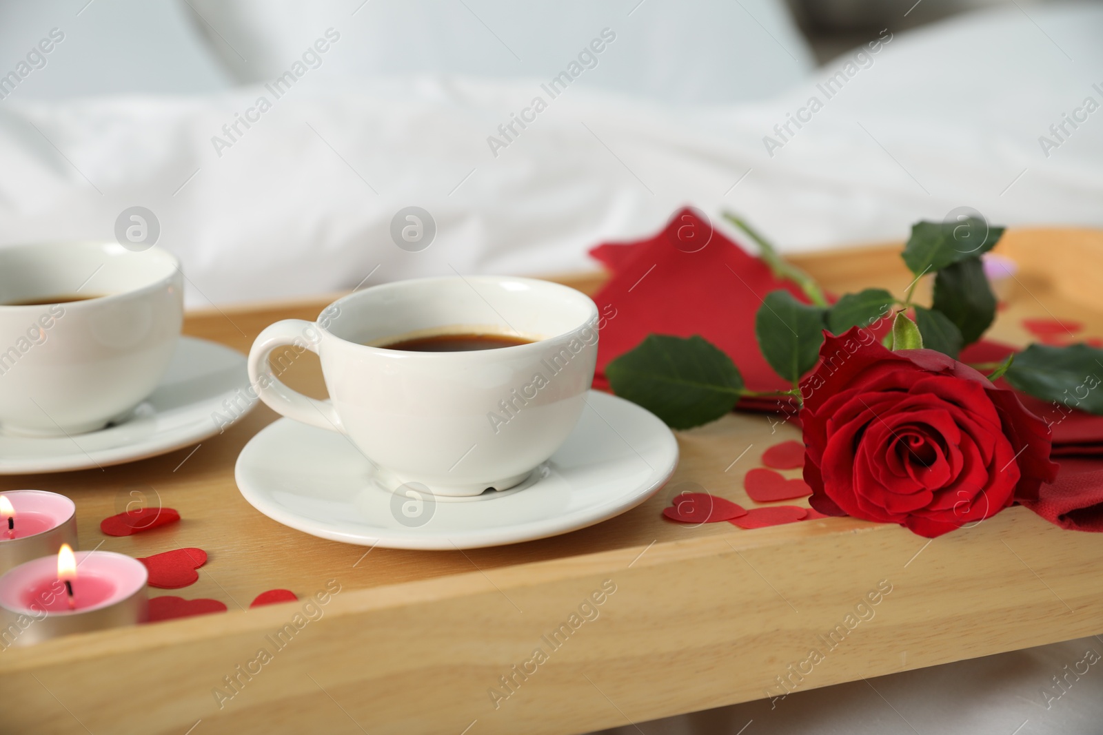 Photo of Wooden tray with burning candles, rose, cups of coffee and red paper hearts on bed indoors, closeup