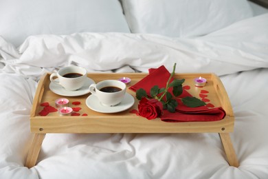 Photo of Wooden tray with burning candles, rose, cups of coffee and red paper hearts on bed indoors