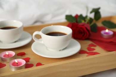 Photo of Wooden tray with burning candles, rose, cups of coffee and red paper hearts on bed indoors, closeup