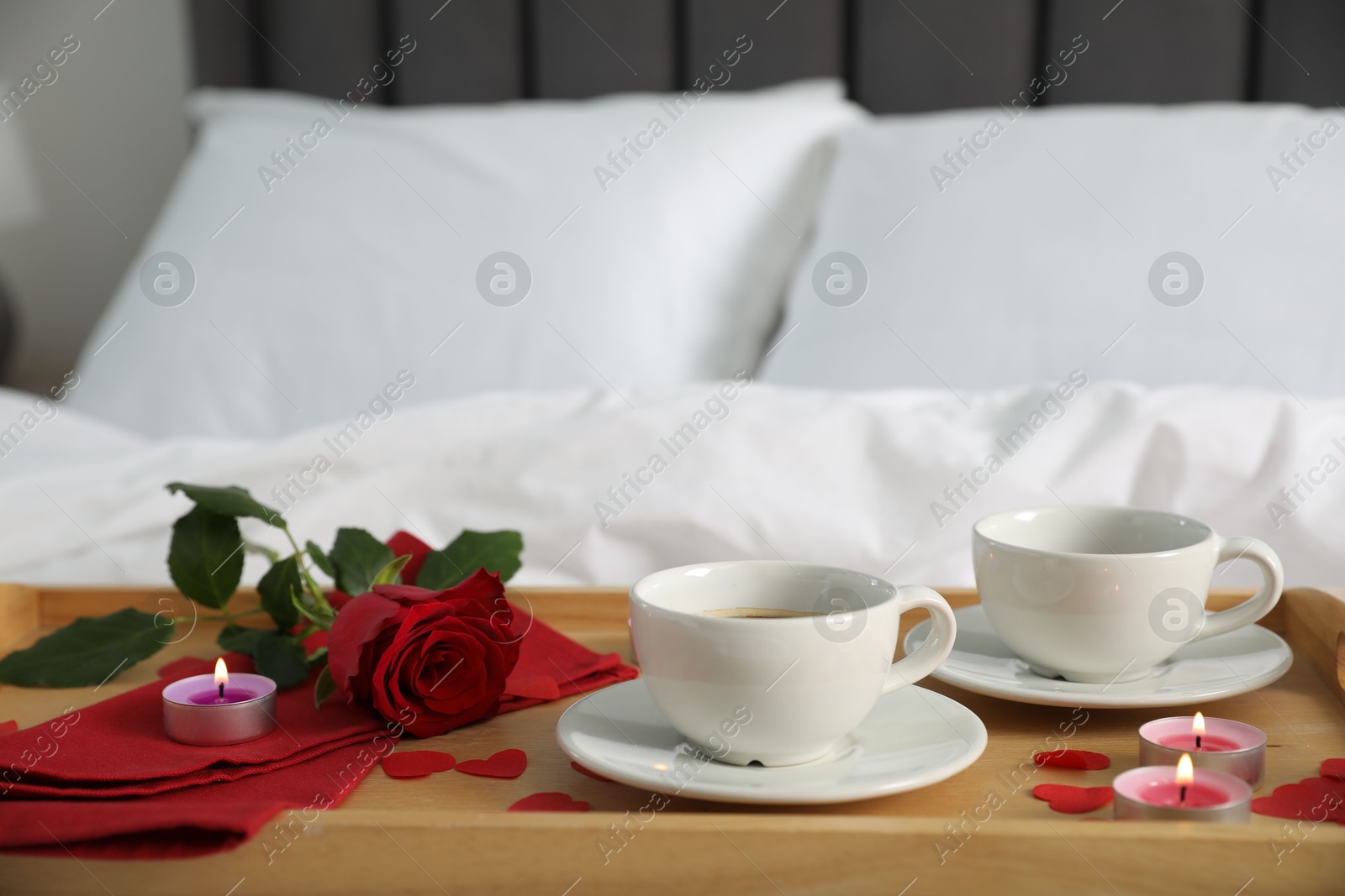 Photo of Wooden tray with burning candles, rose, cups of coffee and red paper hearts on bed indoors