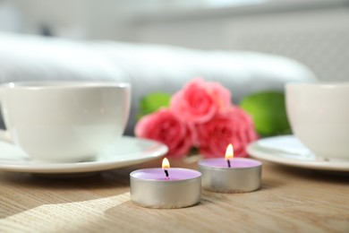 Photo of Burning candles, rose flowers and cups of drink on wooden table indoors