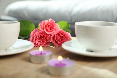 Burning candles, rose flowers and cups of drink on wooden table indoors