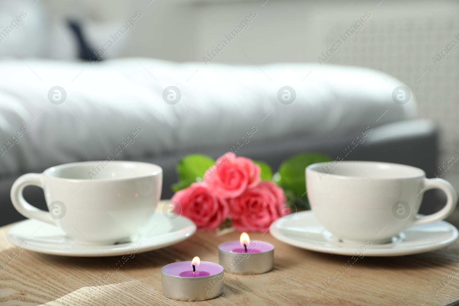 Photo of Burning candles, rose flowers and cups of drink on wooden table indoors