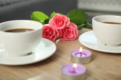 Photo of Burning candles, rose flowers and cups of drink on wooden table indoors, closeup