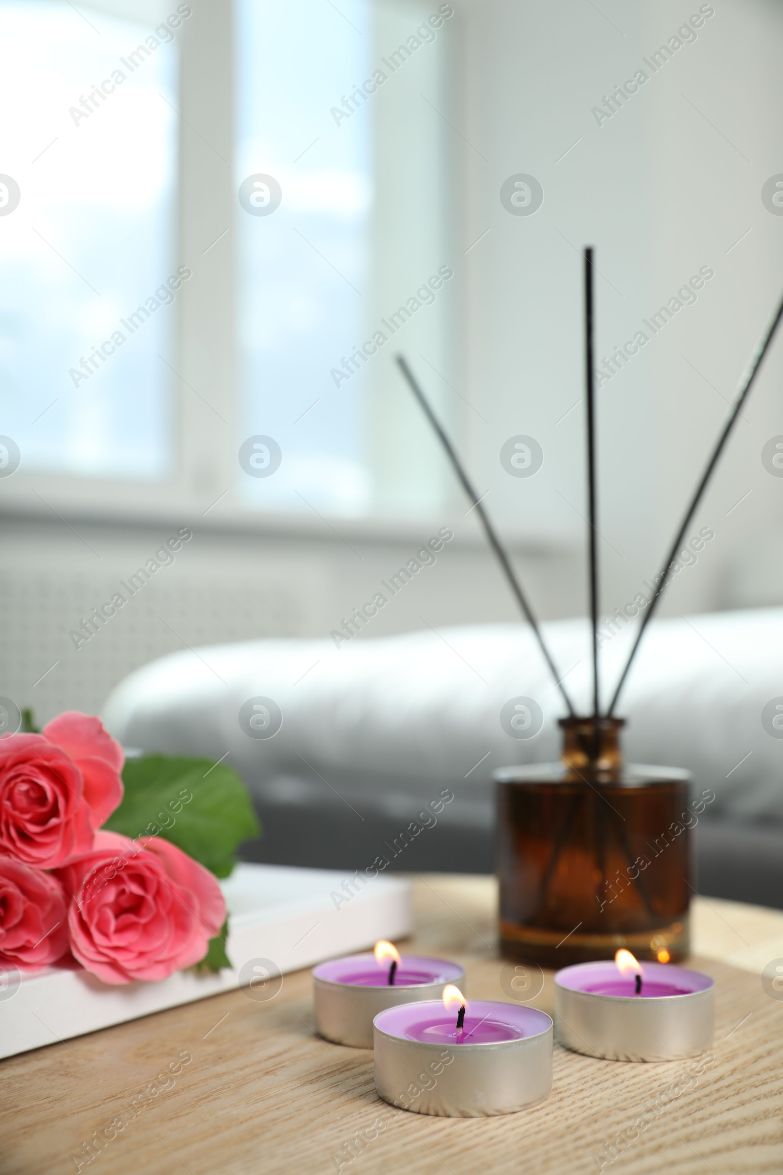 Photo of Burning candles, rose flowers, book and reed diffuser on wooden table indoors