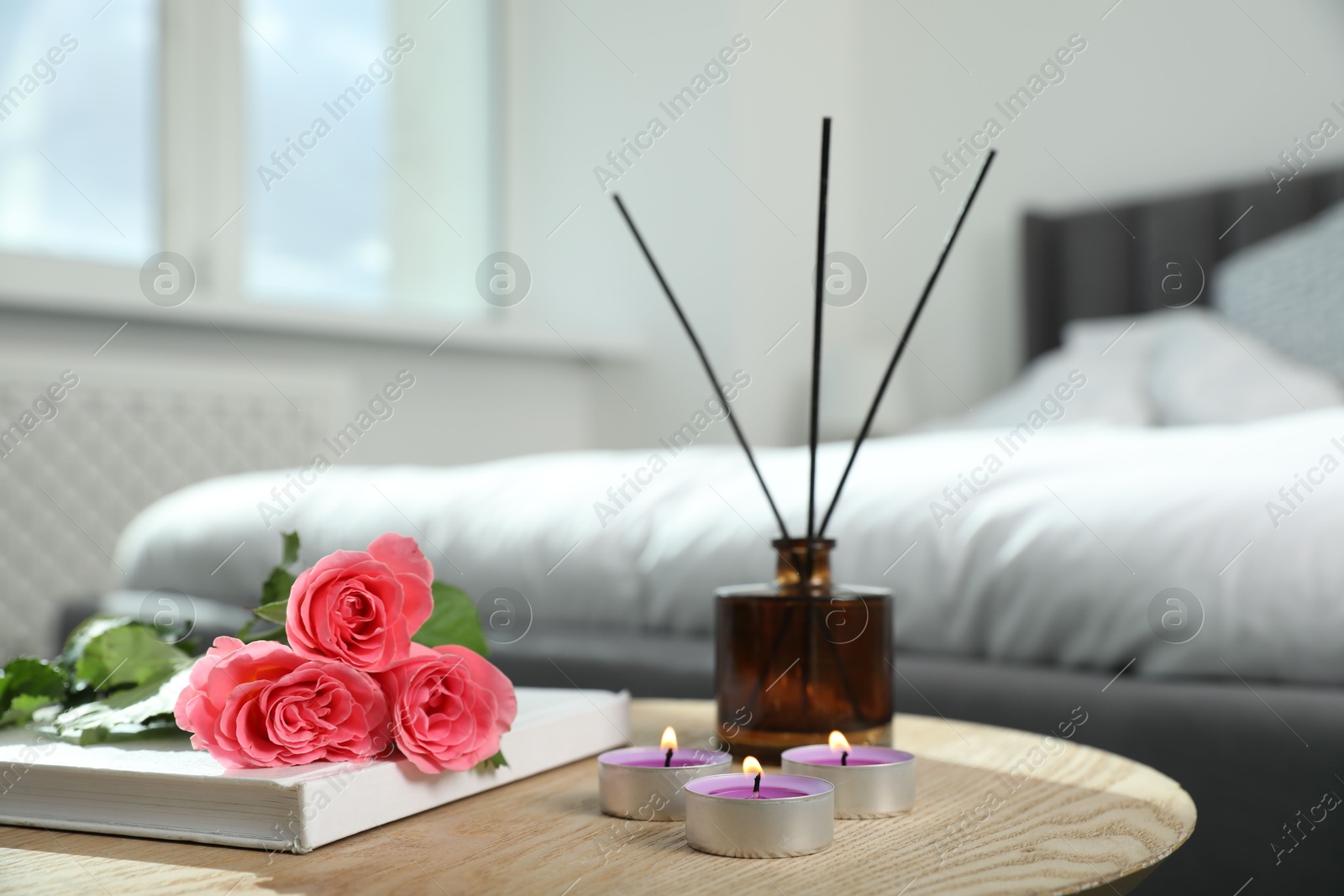 Photo of Burning candles, rose flowers, book and reed diffuser on wooden table indoors